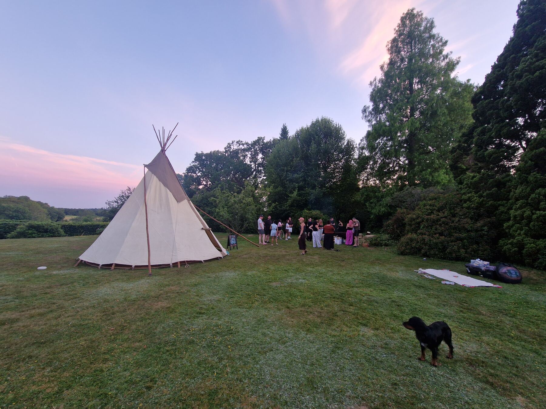 A tipi at dusk with a group gathered beside it under a pink sky