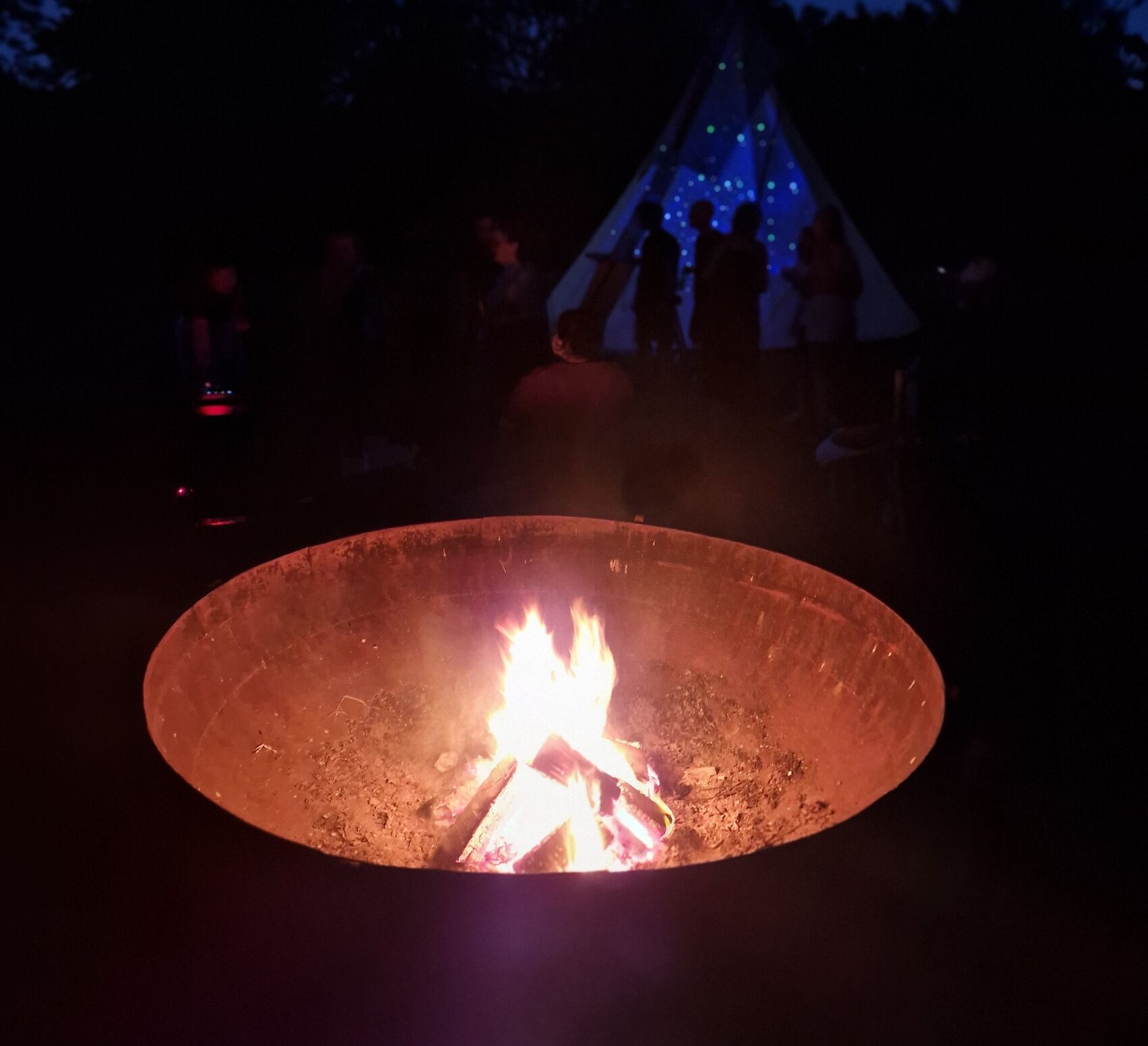 A fire burning in a steel bowl at night, a lit tipi behind it